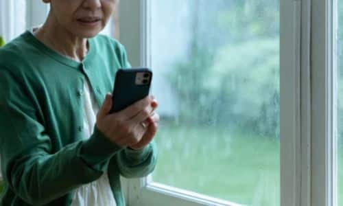 Signora davanti alla finestra di casa, con la finestra bagnata dalla pioggia, riceve un avvisio push delle app mobili